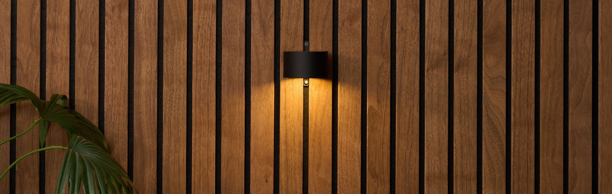 Wall-mounted light fixture on a wooden panel wall with a plant and cushion in the foreground.